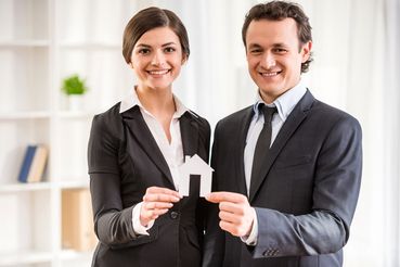 Ein Mann und eine Frau im Anzug die ein Papierhaus in der Hand halten